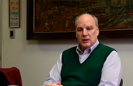 Hombre con suéter verde y camisa de cuadros, sentado en mesa con taza blanca y mantel amarillo.