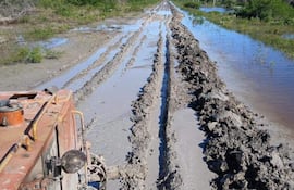 Camino de tierra en malas condiciones con barro y agua al costado, sin personas visibles. Vegetación densa al fondo.