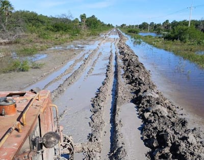 Camino de tierra en malas condiciones con barro y agua al costado, sin personas visibles. Vegetación densa al fondo.
