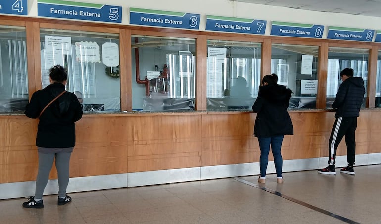 Cuatro personas en la farmacia del IPS, vistiendo ropa casual, se acercan a mostradores numerados en un ambiente simple y funcional.