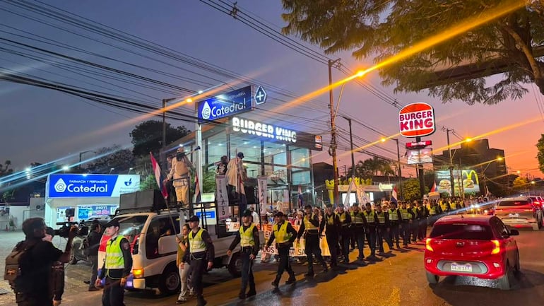 Grupo de médicos en camioneta sosteniendo banderas, mientras policías uniformados organizan la manifestación en un entorno urbano.