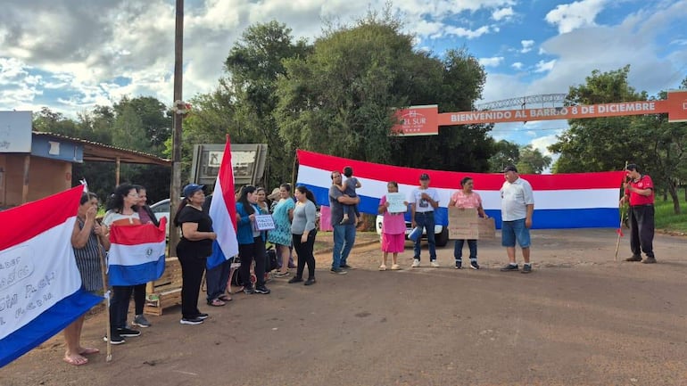 Grupo de manifestantes ondeando banderas de Paraguay en un ambiente exterior, frente a un cartel que dice 'BIENVENIDO AL BARRIO 8 DE DICIEMBRE'.