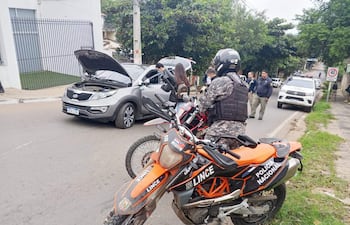 Motocicleta de la Policía Nacional con agente en chaleco táctico y cascos, mientras hombres cerca de un coche gris revisan el motor.