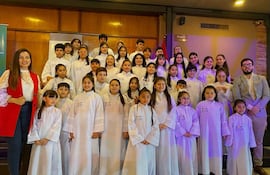 Niños con túnicas blancas sonríen en evento, con adultos sonrientes, en un salón decorado con madera.