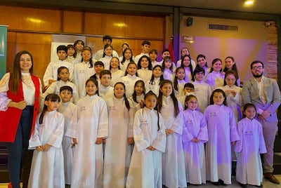 Niños con túnicas blancas sonríen en evento, con adultos sonrientes, en un salón decorado con madera.