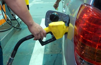 Persona con manguera amarilla de combustible en mano, en una estación de servicio con dispensadores visibles de fondo.