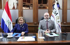 Alicia Pucheta sonríe en traje azul, mientras Enrique Berni, serio en traje claro, lo observa en una reunión formal.