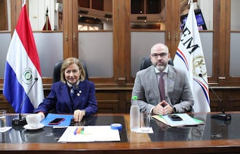 Alicia Pucheta sonríe en traje azul, mientras Enrique Berni, serio en traje claro, lo observa en una reunión formal.