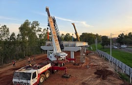 Obreros en el sitio de construcción con grúas levantando vigas, rodeados de maquinaria pesada y vegetación.