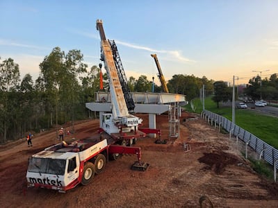 Obreros en el sitio de construcción con grúas levantando vigas, rodeados de maquinaria pesada y vegetación.