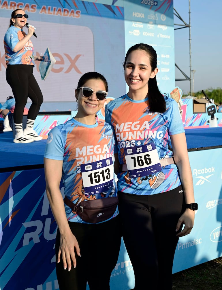 Dos mujeres con camisetas del evento MEGA RUNNING 2026, una lleva gafas de sol, ambas sonriendo en un ambiente exterior.