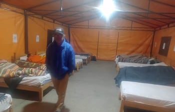 Hombre con abrigo azul y pantalones claros de pie en albergue temporal, rodeado de camas y mantas de colores.