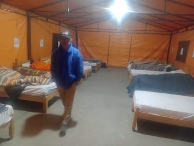 Hombre con abrigo azul y pantalones claros de pie en albergue temporal, rodeado de camas y mantas de colores.