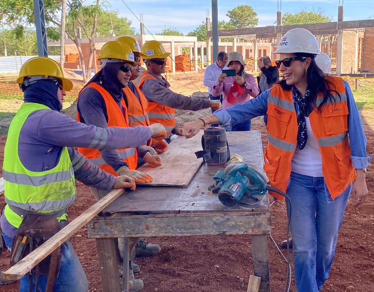 Claudia Centurión sonríe con chaleco naranja y casco blanco mientras saluda a un hombre en casco amarillo en obra.