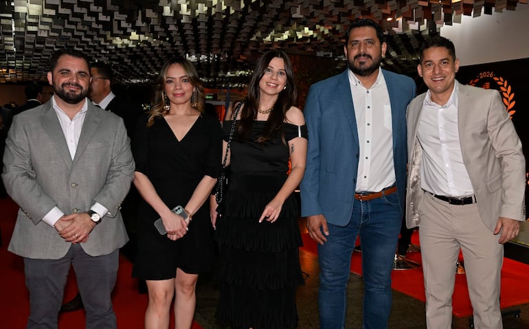 Cinco personas en gala: hombre de traje gris serio, mujer en vestido negro con teléfono, y otros sonriendo en vestido negro y trajes claros.