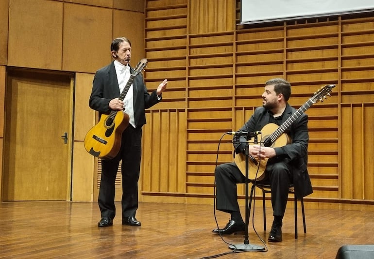 Acosta, en chaqueta oscura y camisa blanca, habla mientras otro guitarrista lo observa, ambos en un auditorio.