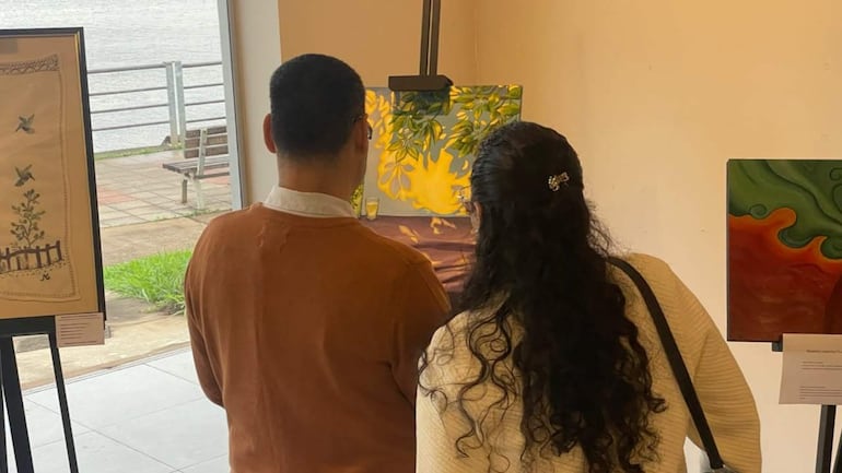 Hombre en suéter marrón y mujer con blusa clara contemplan una pintura en galería de arte, rodeados de obras expuestas.