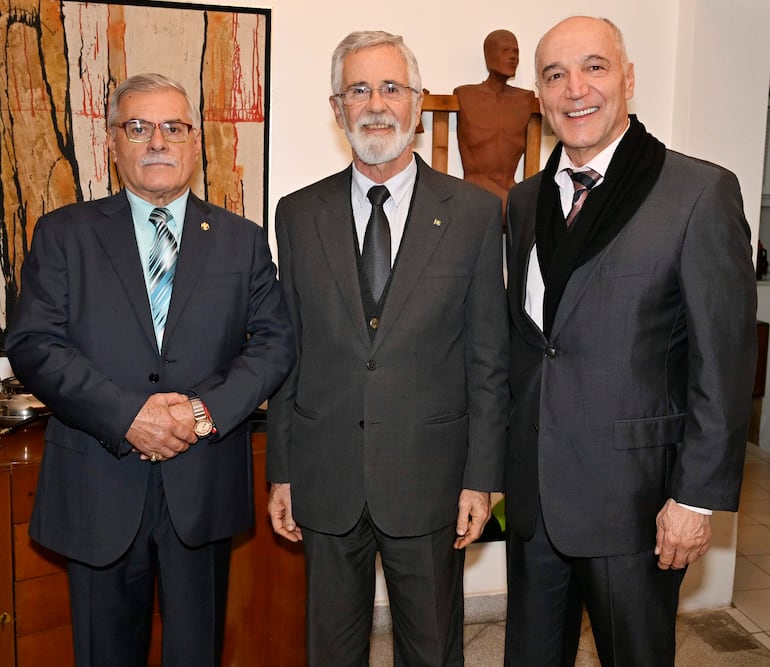 Hombres de pie en trajes formales. Soccoja lleva abrigo negro y sonríe, mientras otros visten trajes oscuros con corbatas. Ambiente decorado.