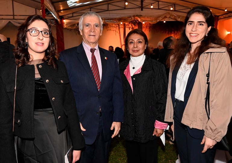 Cuatro personas sonríen y posan en el evento, vistiendo chaquetas elegantes en un ambiente decorado con luces.