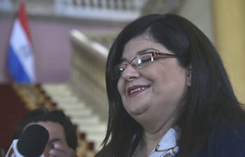 Carolina Llanes sonriendo y usando gafas, mientras responde preguntas en un ambiente oficial con bandera paraguaya de fondo.