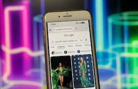 Pantalla de teléfono móvil mostrando búsqueda de Jennifer Lopez en vestido verde Versace, con fondo colorido.