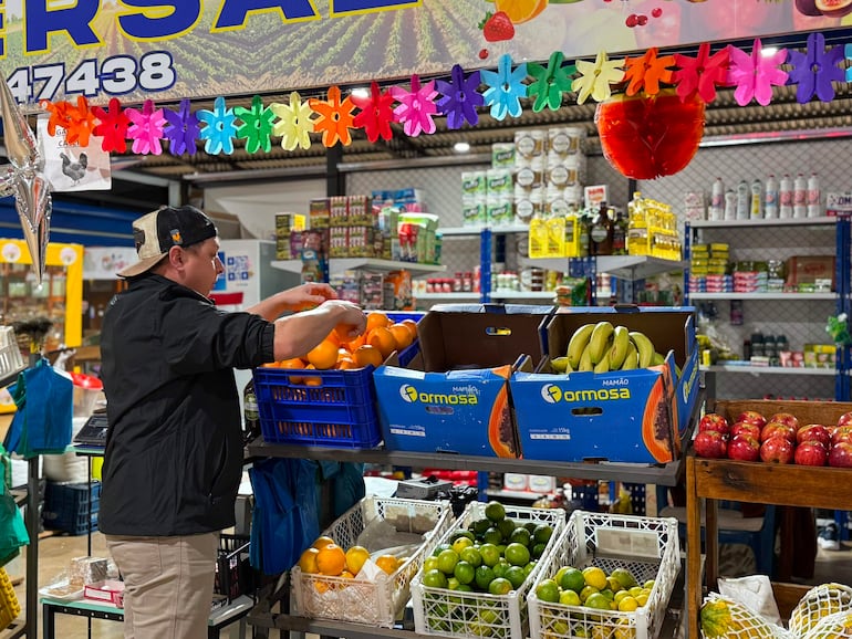 Hombre de espaldas en camiseta negra y gorra organiza naranjas, rodeado de frutas y decoraciones coloridas en un mercado.