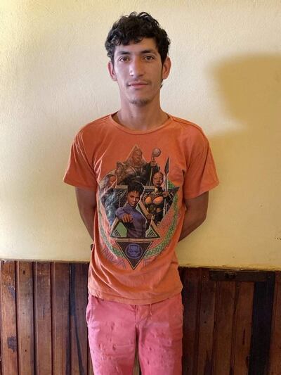 Hombre de pie en ambiente interior, lleva camiseta naranja con diseños gráficos y pantalones rosa, fondo claro y estructura de madera.
