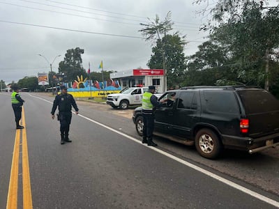 Dos policías con uniforme oscuro y chalecos reflectantes conversan con un conductor en un vehículo negro en una carretera nublada.
