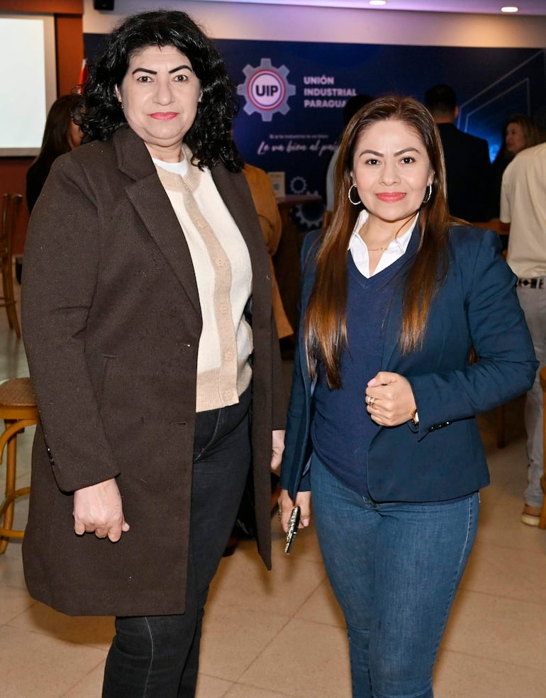 Dos mujeres sonrientes, una con abrigo marrón y otra con chaqueta azul, posan en un evento de la UIP con logotipo visible en el fondo.