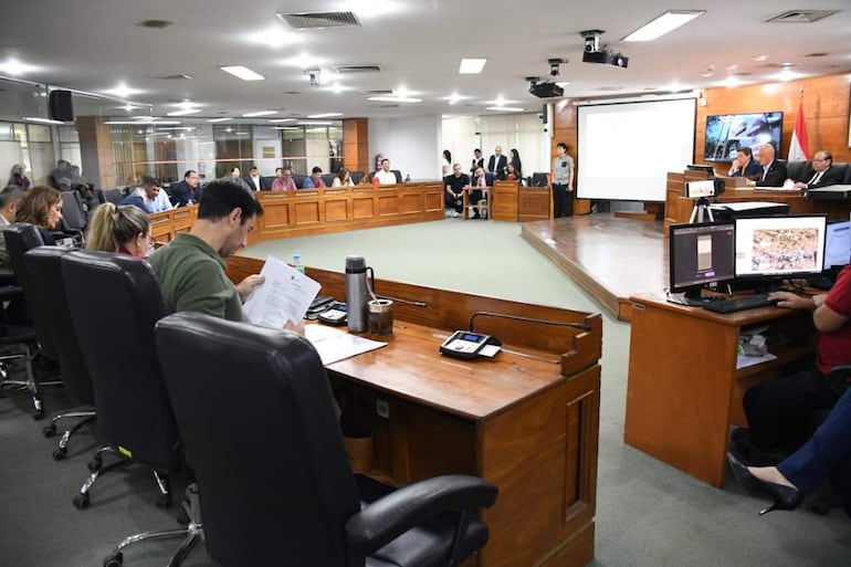 Asistentes en salón formal discutiendo, con Carlos Reyes presente en mesa principal, mientras algunos toman notas y prestan atención.