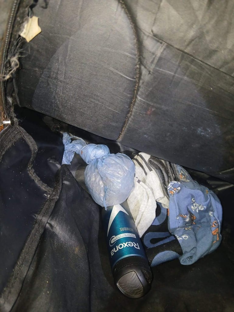 Interior de un bolso oscuro con aerosol Rexona, ropa y un paquete azul. Sin personas visibles.
