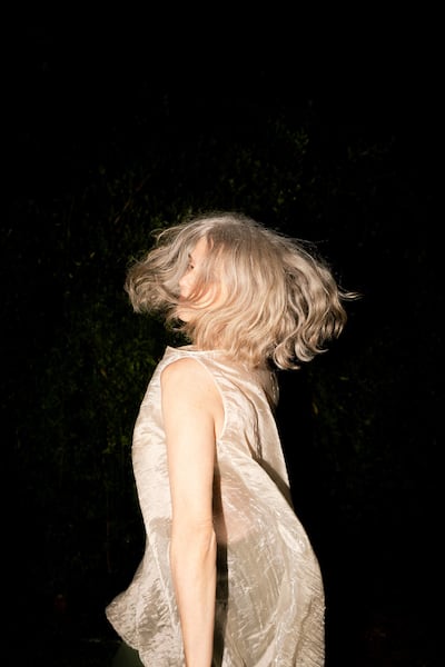Mujer de cabello canoso gira, vistiendo blusa clara que brilla, en un entorno oscuro que evoca un ambiente artístico.