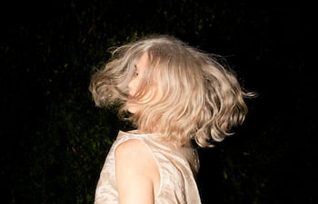 Mujer de cabello canoso gira, vistiendo blusa clara que brilla, en un entorno oscuro que evoca un ambiente artístico.