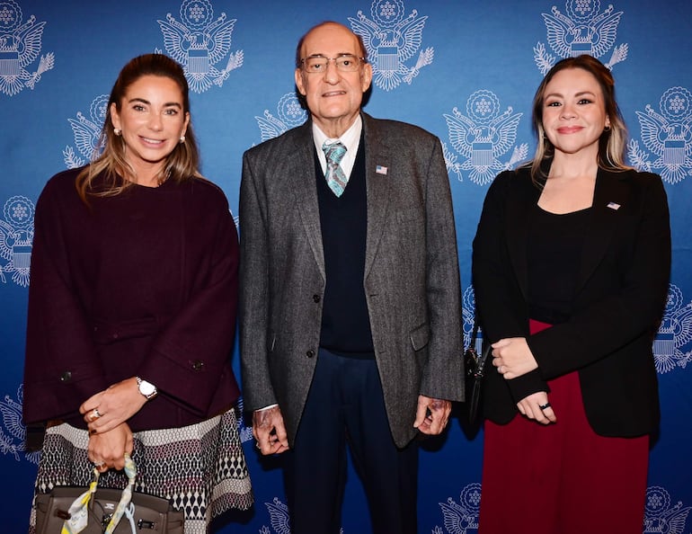 Tres personas sonriendo en un evento, un hombre de traje gris en el centro, flanqueado por dos mujeres, una con abrigo oscuro y otra en blazer negro y falda roja.