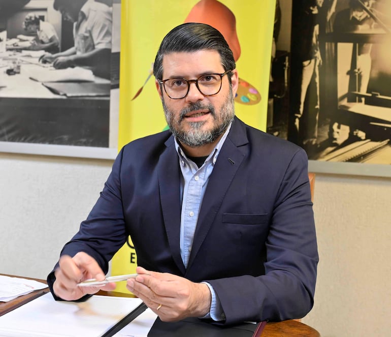 Hombre con barba y cabello oscuro, vestido con traje oscuro y camisa clara, sentado en una oficina sosteniendo un bolígrafo.