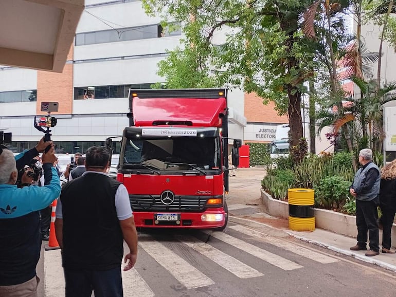 Camión rojo de justicia electoral estacionado con periodistas observando y tomando fotos en un entorno urbano.