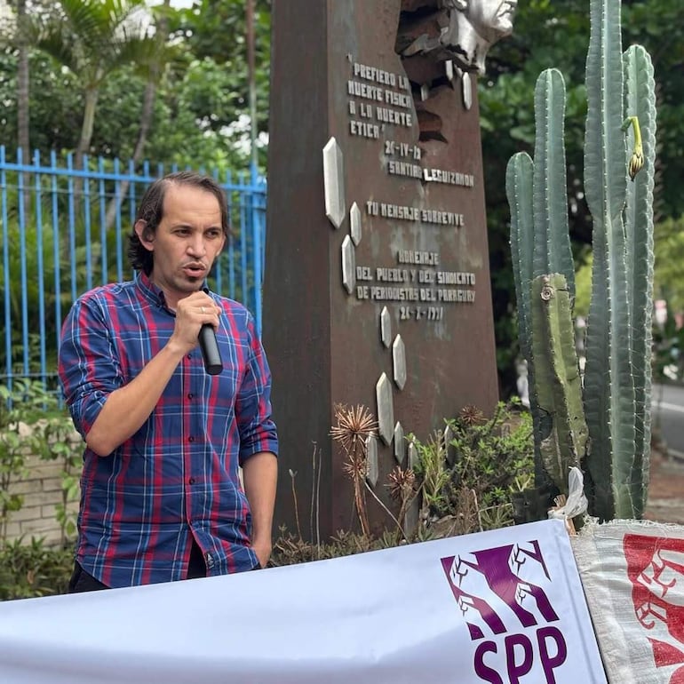 Hombre con camisa de cuadros habla frente a un monumento, rodeado de vegetación y una pancarta del SPP.