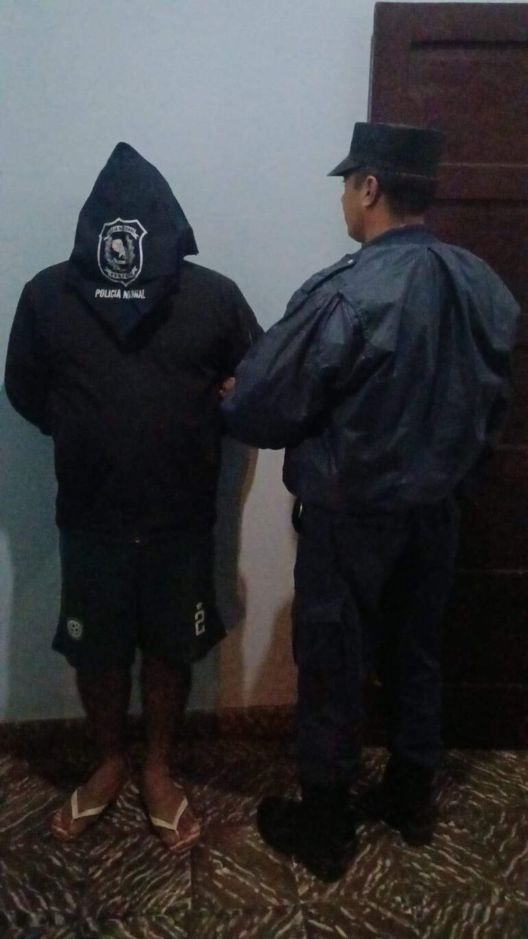 Hombre con sudadera negra y pantalones cortos bajo custodia de un oficial de policía en uniforme azul oscuro.