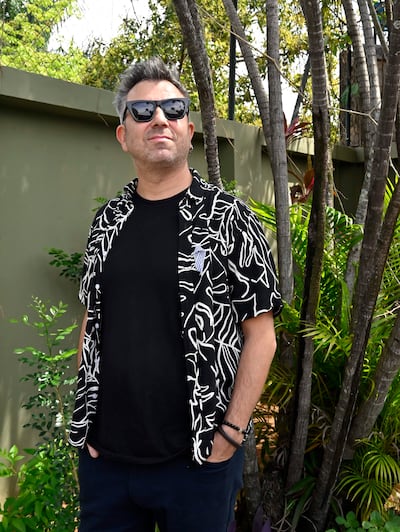 Hombre de pie en entorno natural, con camisa de estampado negro y blanco, y gafas de sol, posando relajado ante una pared verde.