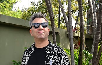 Hombre de pie en entorno natural, con camisa de estampado negro y blanco, y gafas de sol, posando relajado ante una pared verde.