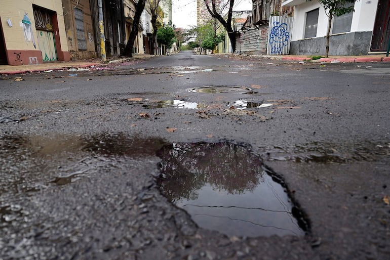 Calle vacía en Asunción con charcos reflejando árboles y edificios, pavimento agrietado y hojas secas.