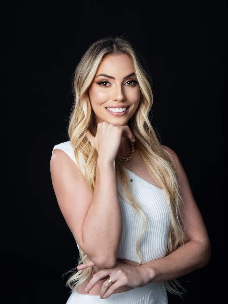Thalía Kehl, mujer rubia con blusa blanca, sonríe mientras posa con confianza en un fondo negro.