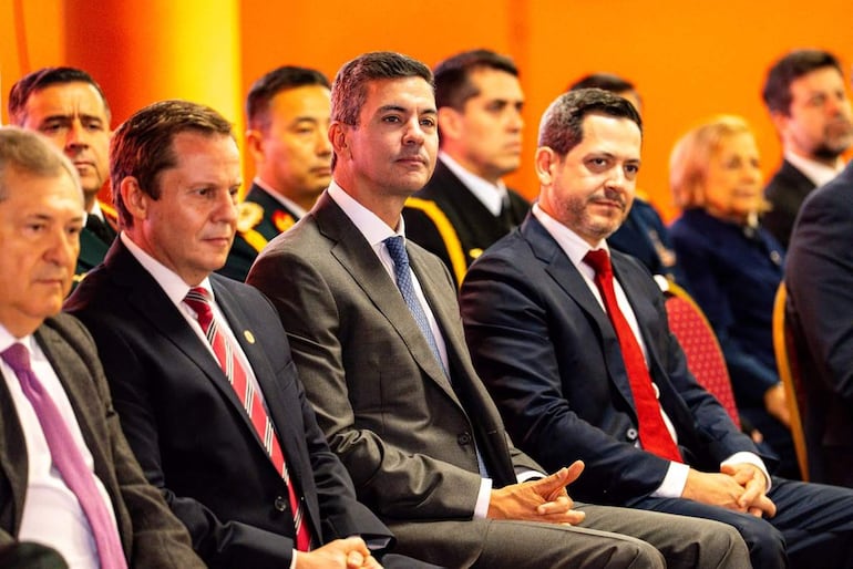 Santiago Peña en chaqueta gris y corbata púrpura, junto a hombres en trajes oscuros y una mujer rubia, en un auditorio formal.