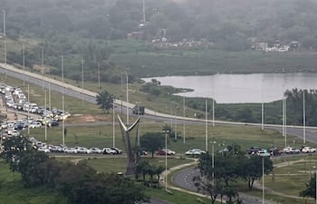 Extensa fila de vehículos paralizados en la Costanera, con un monumento abstracto al frente y árboles al fondo.