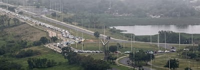 Extensa fila de vehículos paralizados en la Costanera, con un monumento abstracto al frente y árboles al fondo.