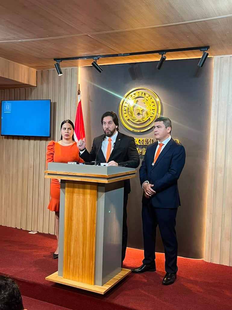 Rubén Velázquez en podio, gesto de oratoria; Leidy Galeano en vestido naranja y José Oviedo en traje oscuro, atentos.