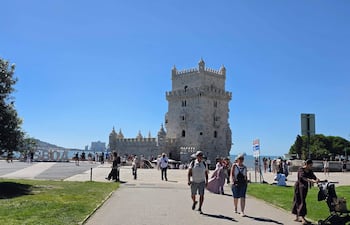 Varias personas con ropa de verano caminan por un paseo peatonal, observando la Torre de Belém bajo un cielo despejado.