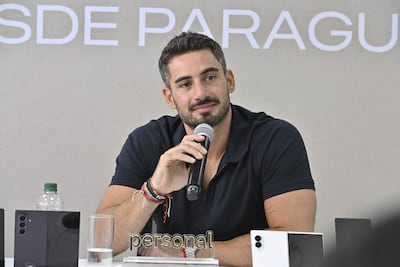 Hombre con camiseta oscura y barba, sentado ante una mesa con micrófono, vaso y teléfonos, hablando en un ambiente luminoso.