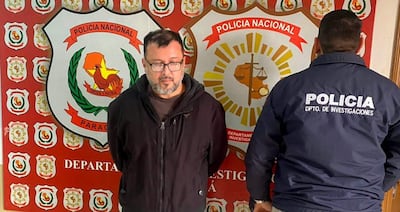 Dos hombres en una comisaría: uno de pie con chaqueta oscura, otro de espaldas con chaqueta de policía, fondo con escudo policial.