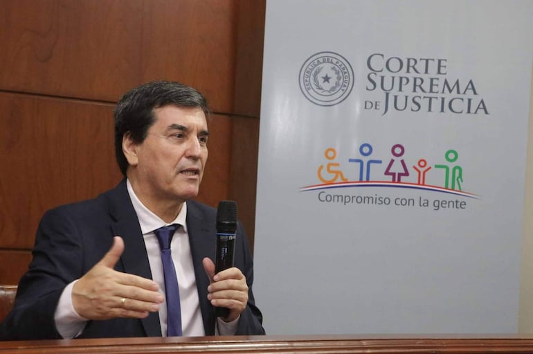 Gustavo Santander Dans vestido formalmente, sosteniendo un micrófono y hablando ante un panel que dice 'Corte Suprema de Justicia'.